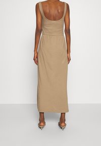 Žena stojící zády, oblečená v bezramínkových béžových maxi šatech a zlatých sandálech s nízkým podpatkem na jednobarevném pozadí.