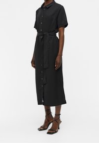 Robe noire boutonnée devant avec manches courtes, ceinture nouée à la taille et coupe droite, réalisée dans un tissu léger. Portée avec des sandales noires à fines lanières.