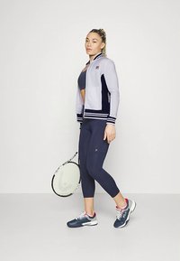 FILA sportsjakke i lys grå med marineblå detaljer, glidelås foran, stripete mansjetter. Kombinert med marineblå leggings og tennissko, holder en racket.