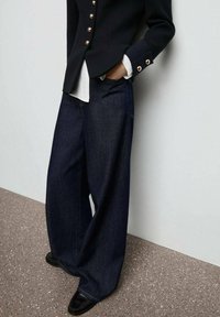 Široké nohavice z tmavého denimu s hladkou texturou, které mají hluboké kapsy a volný střih, v kombinaci s černým sakem a bílou košilí.