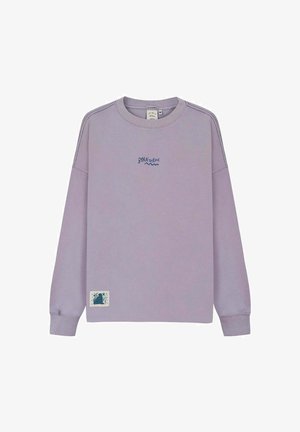 Blondynka w jasnofioletowej bluzie z kapturem z niebiesko-zielonym tygrysem i napisem "Born to Roar" na plecach, stojąca na piaszczystym podłożu.