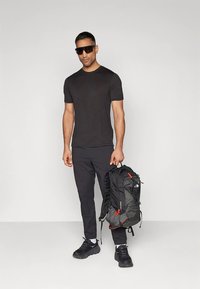 Melnas īsroku krekls, melnas bikses, saulesbrilles un melni ikdienas apavi. Viņš tur pelēku un melnu mugursomu ar oranžām detaļām.