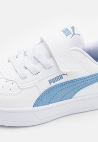 Zapatilla blanca con acentos en azul claro, presenta una correa de velcro, parte superior de cuero texturizado y diseño perforado en la zona de los dedos.