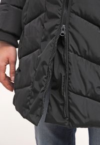 Chaqueta de plumas negra con un diseño acolchado, que presenta un cierre frontal y puños acanalados. La textura parece suave y brillante.