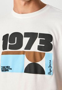 Hvid skjorte med fed sort tekst "1973", geometriske former i brun, sort og blå samt sætningerne "DEN NYE EVOLUTION BEGYNDER" og "Pepe Jeans."