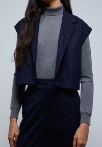 Lyhyempi tummansinivalkoinen raitaliivi harmaan poolopaidan päällä, jossa on muotoiltu design ja pehmeä rakenne. Kainaloaukot ovat leveät ja avoimet.