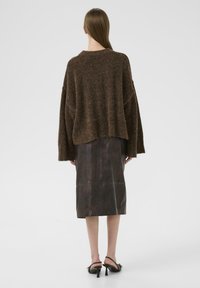 Rumahtangan cokelat oversized terbuat dari bahan lembut dan bertekstur, dipadukan dengan rok kulit cokelat tua sepanjang sedang dan sandal bertumit hitam.