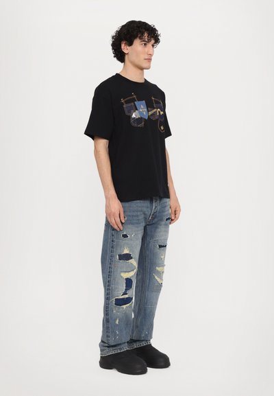 T-shirt noir à manches courtes avec un design graphique coloré à l'avant, associé à un jean en denim bleu déchiré et des chaussures noires.
