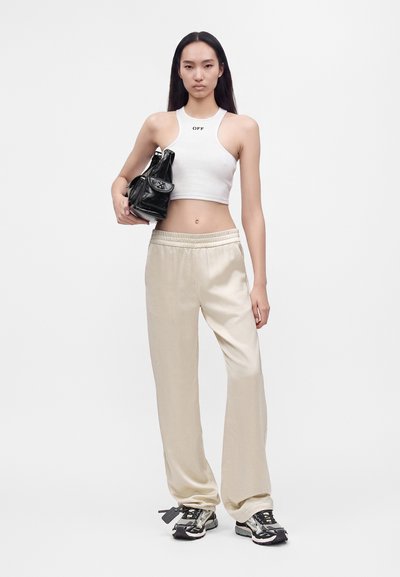Jeune femme en débardeur court blanc, pantalon ample crème, baskets noires et blanches, tenant un petit sac à main noir sous le bras.