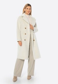 Abrigo exterior de mezcla de lana blanca con diseño de doble botonadura, gran cuello y detalles en botones marrones. La modelo lleva pantalones y zapatos beiges.