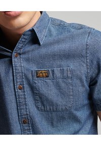 Koszula jeansowa z krótkim rękawem w średnim odcieniu niebieskiego, z kieszenią ozdobioną czarną i złotą metką marki oraz brązowymi guzikami na listwie.