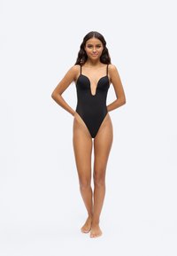 Frau steht barfuß da, trägt einen schwarzen Einteiler mit dünnen Trägern und tiefem V-Ausschnitt, lächelt und hat die Hände hinter dem Rücken verschränkt.