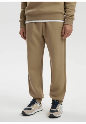 Mężczyzna ubrany w beżowe spodnie dresowe, jasnobrązową bluzę, białą koszulę oraz beżowo-granatowe sneakersy, stojący na białej podłodze.