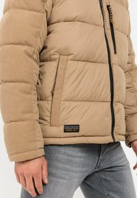 Tan Puffer-Jacke mit strukturierter Oberfläche, horizontalen Nähten, Reißverschlusstaschen und einem Etikett mit "MADE FOR DAILY ADVENTURES SINCE 1977".