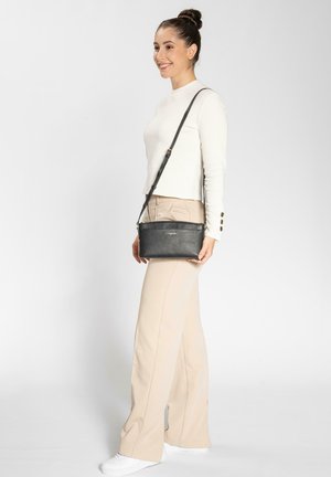 Borsa a tracolla in pelle nera textured con cerniera, design slim, tracolla regolabile e branding sottile. Indossata da una donna in abbigliamento chiaro.