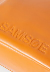 Oranžna usnjena torba z odtisnjenim logotipom "SAMSOE", ki se ponaša z gladko teksturo in čistim, pravokotnim oblikovanjem.