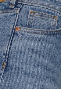 Tissu en denim bleu clair avec une poche visible, des rivets en cuivre et des surpiqûres orange. La texture est légèrement usée, typique du denim.