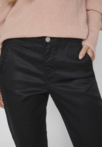 Pantalones negros con una textura suave, que cuentan con bolsillos delanteros y un cierre de botón plateado. Combinados con un suéter de punto acanalado en rosa claro.