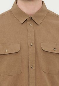 Καφέ πουκάμισο με κουμπιά και δύο μπροστινές τσέπες με καπάκι, με κλασικό γιακά, που φορά ένα άτομο.