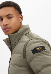 Jonge man in een olijfgroene gewatteerde jas met een PME Legend-logo op de bovenarm, staand tegen een witte achtergrond.