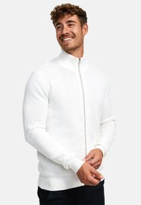Weißer Reißverschluss-Pullover mit einem Zickzackmuster, gerippten Ärmelbündchen und Saum sowie hohem Kragen, aus einem strukturierten Strickmaterial.