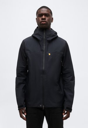 Noor mees, kes kannab musta kapuutsiga jopet esist lukuga ja väikese kollase logoga rinnal, seisab ühevärvilise tausta ees.