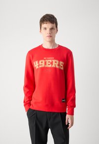 Sweatshirt vermelho com "49ERS" e "SAN FRANCISCO" em impressão dourada. Feito de material semelhante a fleece, com punhos e bainha canelados. Etiqueta de logotipo preta.