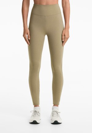 Vrouw in hoogsluitende, beige textuur leggings en witte sneakers, staand tegen een effen witte achtergrond.
