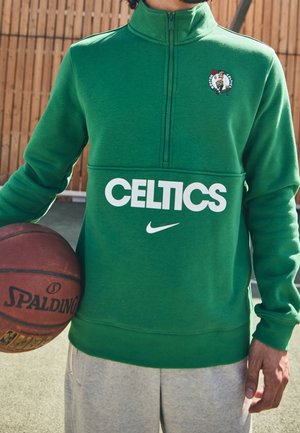 Osoba w zielonej bluzie Boston Celtics trzymająca piłkę do koszykówki Spalding na tle drewnianego ogrodzenia.