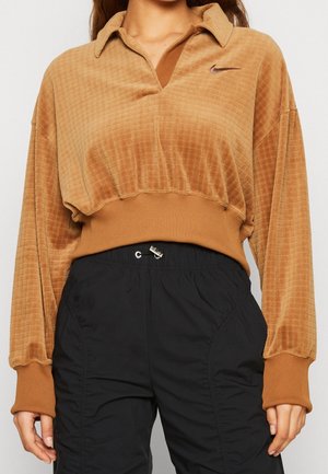 Personne portant un sweat-shirt court marron texturé avec col et logo Nike, associé à un pantalon à taille élastique noir.