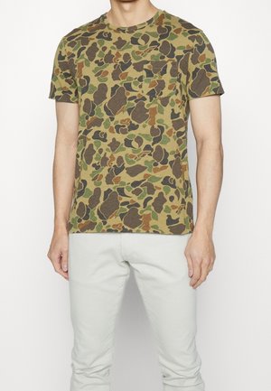 Mann trägt kurzärmeliges T-Shirt mit grün-braunem Tarnmuster und Brusttasche sowie hellen Hosen, steht vor einfarbigem Hintergrund.