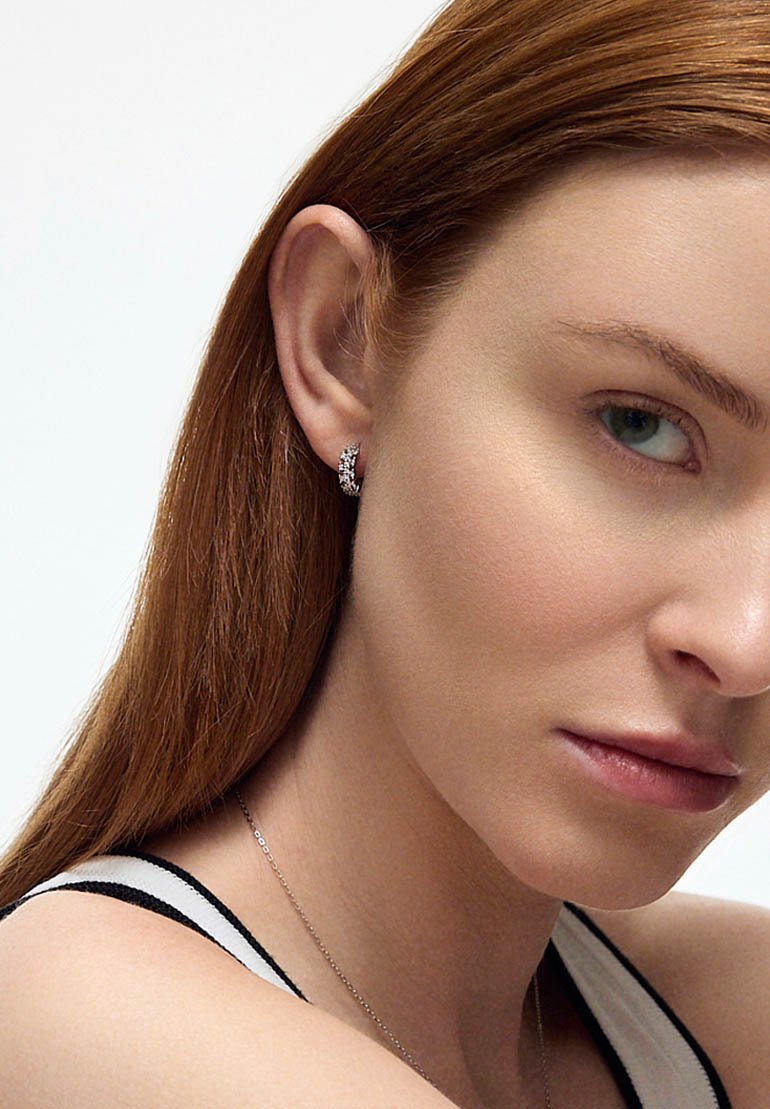 Srebrna kolczyk w formie obręczy z małymi, osadzonymi przezroczystymi kamieniami szlachetnymi; gładka powierzchnia; zaokrąglony kształt. Włosy proste i kasztanowe, oprawiają twarz.