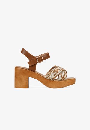 Sandal de tacón cuadrado de cuero marrón con parte superior de paja trenzada. Cuenta con una correa para el tobillo ajustable con hebilla y textura de madera en la suela.