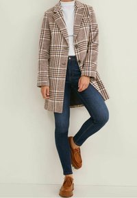 Abrigo largo de cuadros marrones con un diseño entallado, cierre de botones y bolsillos laterales, usado sobre una blusa blanca y jeans ajustados azules con zapatos marrones.