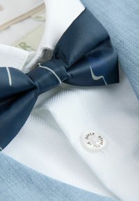 Tamsiai mėlynas satino peteliškė su baltais dryželiais, derinama su balta tekstūruota marškinių palaidine ir šviesiai mėlynu švarku; antsegis pažymėtas užrašu „TED BAKER.“