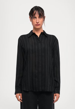 Donna che indossa una camicia nera a maniche lunghe con bottoni e sottili righe bianche verticali, in piedi contro uno sfondo leggero e semplice.
