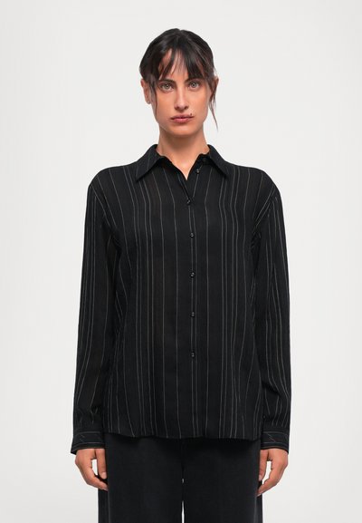 Donna che indossa una camicia nera a maniche lunghe con bottoni e sottili righe bianche verticali, in piedi contro uno sfondo leggero e semplice.