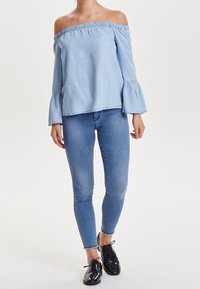 Lyseblå off-shoulder top med rynket halsudskæring og lange klokkeærmer, parret med tætsiddende blå jeans og sorte patenterede sko.