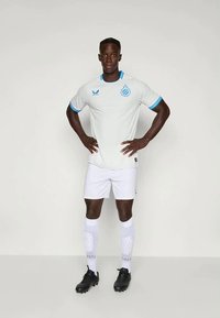 Camisola de futebol branca de manga curta com detalhes azuis, apresentando o emblema do clube. Combinada com calções brancos e meias até ao joelho. Chuteiras de futebol pretas.