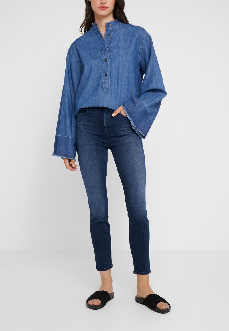 Camisa de mezclilla azul con cuello alto, tapeta de botones y mangas anchas. Combinada con vaqueros ajustados azules oscuros y sandalias negras tipo slides.