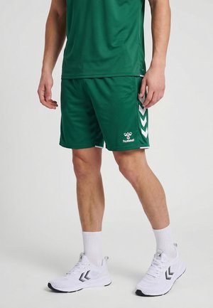 Man draagt donker groene sportshorts en bijpassend shirt, witte sportsokken en witte sportschoenen met zwarte logodetails.