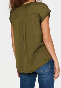 Olijfgroene blouse met korte mouwen, subtiel stippenpatroon, gebogen onderkant en een ontspannen pasvorm. De achterkant heeft een yoke en een centrale plooi.