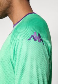 Zelena atletska majica z vzorcem teksture, s vijoličastim ovratnikom in vijoličastim logotipom na levem rami.