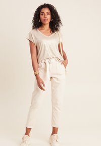 Beige metallic v-hals t-shirt met korte mouwen, gecombineerd met lichte crèmekleurige jogpants met koord. Model draagt casual sneakers en gouden accessoires.