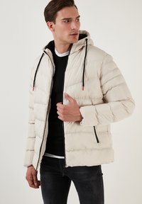 Jachetă puffer bej deschis cu quilt horizontal, fermoar negru și glugă ajustabilă. Prezintă o textură netedă și manșete elastice.