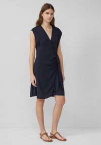 Marineblaue Wickelkleid mit kurzen Ärmeln, tiefem V-Ausschnitt und gerafftem Seitenelement. Tragend mit braunen Schnürsandalen vor einem neutralen Hintergrund.