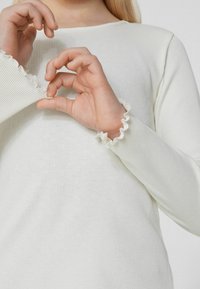 Długi rękaw, prążkowany top z miękkiego białego materiału, z falbaniastymi mankietami. Na zdjęciu widać ręce dostosowujące mankiet, aby lepiej pokazać szczegóły.