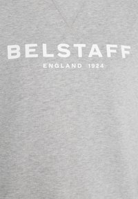 Tissu gris avec texte blanc « BELSTAFF » au-dessus d'un texte plus petit « ENGLAND 1924 » centré sur le devant, probablement un sweat-shirt ou un hoodie.