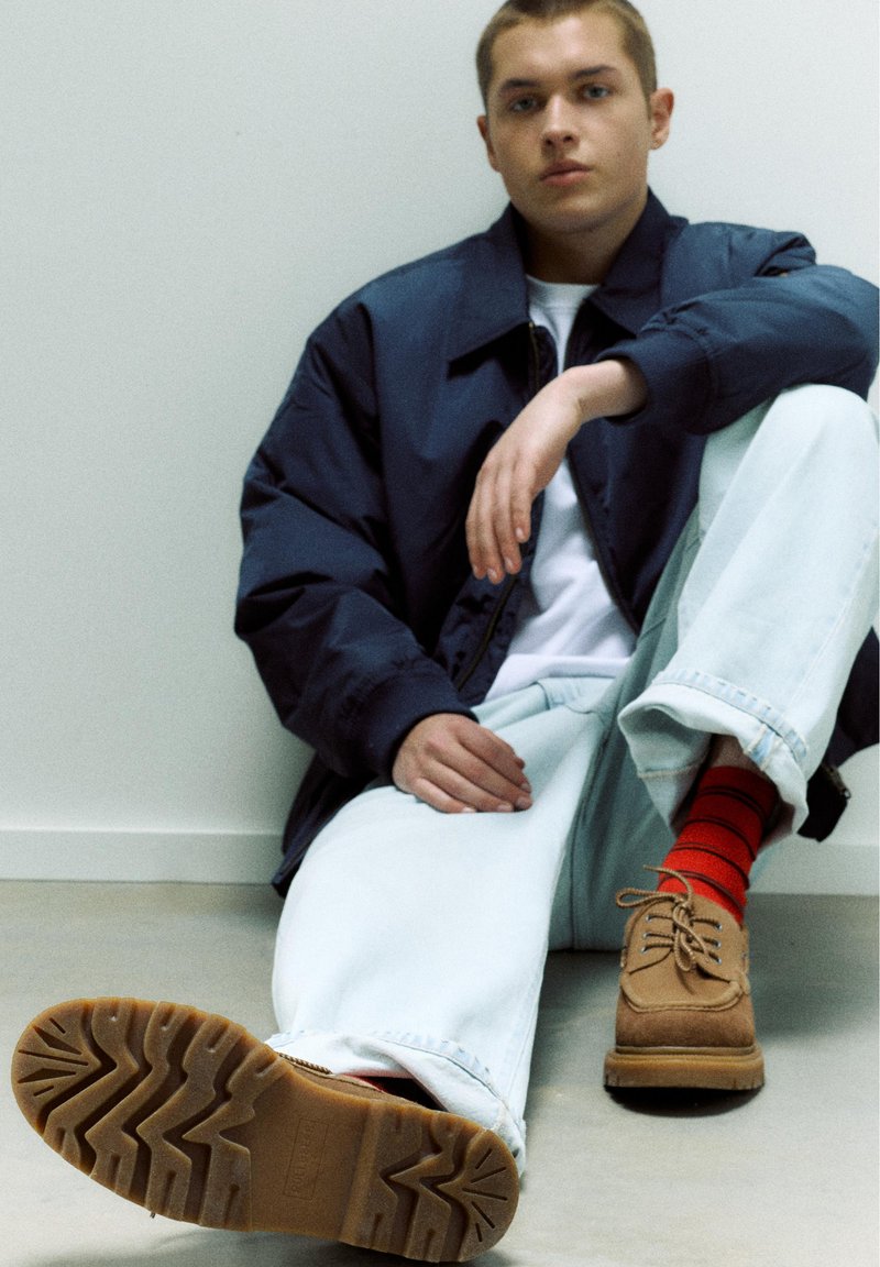 Jeune homme assis par terre, portant une veste bleu marine, un pantalon blanc, des chaussettes rouges rayées et des chaussures marron à lacets, regardant devant avec une expression neutre.
