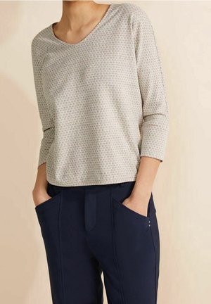 Vrouw draagt een lichtgrijze, met patroon bedrukte top met lange mouwen en een ronde hals, gecombineerd met donkerblauwe broek, met handen in de zakken.